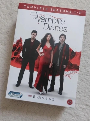 The Vampire Diaries säsong 1-3 box - Box med The Vampire Diaries säsong 1-3 på DVD. Omslaget är vitt med röd och svart design och visar tre karaktärer. Innehåller totalt 15 skivor. Perfekt för dig som gillar vampyrserier och drama.