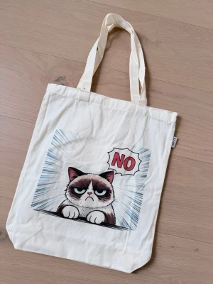 Tygkasse med Grumpy Cat-tryck - Cool shoppingväska i naturvitt tyg med ett stort tryck av Grumpy Cat och texten 'NO' i serietidningsstil. Väskan har långa handtag och är perfekt för att bära böcker, mat eller annat du behöver. Materialet är bomull och designen är både rolig och unik.