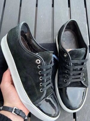 Svarta sneakers från Lanvin - Snygga svarta sneakers från Lanvin med ovandel i mocka och lackad tå. Skorna har svarta skosnören, vita sulor och metallfärgade öljetter. Perfekta för dig som gillar en stilren och lyxig look med en mix av material.