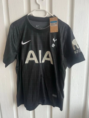 Tottenham Hotspur svart matchtröja Nike - Svart Tottenham Hotspur matchtröja från Nike med diskret rutmönster, vita loggor och tryck. Stort AIA-tryck på bröstet, klubbmärke och Nike-logga. Nummer 15 och namnet Bergvall på ryggen. Korta ärmar och sportig passform.