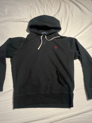 Svart hoodie från Polo Ralph Lauren - Svart hoodie från Polo Ralph Lauren med klassisk röd broderad logga på bröstet. Hoodien har vit dragsko, känguruficka och mjukt material som känns skönt mot huden. Perfekt för en avslappnad och stilren look.