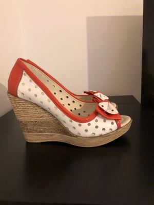 Prickiga kilklackade äkta läder pumps med rosett - Snygga pumps med kilklack i repmaterial, vita med röda prickar och detaljer. Öppen tå och dekorativ rosett framtill. Perfekt för dig som vill sticka ut med en retroinspirerad look. Skorna har en lekfull design och är tillverkade i syntetmaterial.
