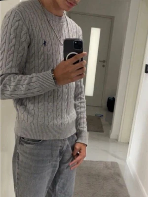 Grå kabelstickad tröja Polo Ralph Lauren - Snygg grå kabelstickad tröja från Polo Ralph Lauren med rund halsringning och klassisk broderad logga på bröstet. Tröjan har ribbade muddar och är perfekt för lager-på-lager-stil. Materialet är mjukt och bekvämt. Nypris: 1946 kr (Zalando)