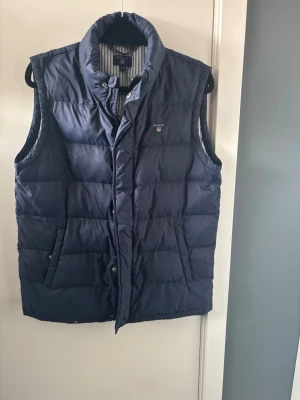 Mörkblå dunväst från Gant - Snygg mörkblå dunväst från Gant med ståkrage och två fickor framtill. Västen har quiltad design och stängs med dragkedja och tryckknappar. Perfekt för lager-på-lager och har klassisk Gant-logga på bröstet.