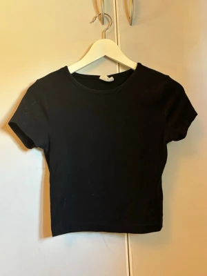Svart croppad t-shirt - En enkel svart croppad t-shirt med rund hals och korta ärmar. Perfekt basplagg som funkar till det mesta och är lätt att matcha. T-shirten är i mjukt material och har en tight passform.