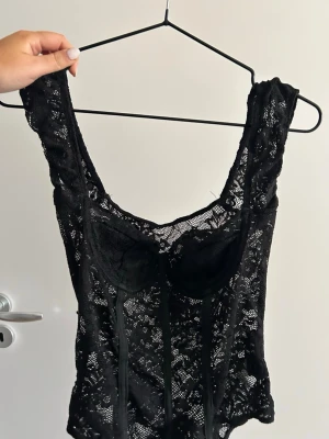 Svart spetsbody - Säljer en svart body i spets med en urringning och markerade kupor som har skydd i. Bodyn har breda axelband och är helt täckt av blommig spets, vilket ger en snygg look, passar perfekt till jeans eller kjol. 