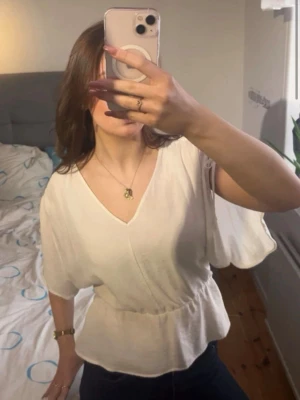 Vit v-ringad blus med resårmidja - Snygg vit blus med v-ringning och korta, vida ärmar. Toppen har resår i midjan som ger en fin form och ett luftigt fall. Perfekt till jeans eller kjol för en fräsch look.