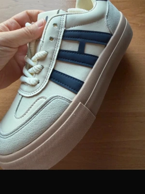 Beige sneakers med blå detaljer 38 - Snygga sneakers i beige med svart ränder på sidorna och tjock beige sula. Skorna har vita snören och detaljer i mockaimitation framtill och baktill. Perfekta för dig som gillar en stilren och sportig look.