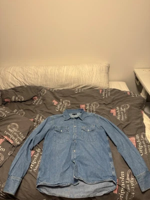 Blå jeansskjorta från GANT - Snygg blå jeansskjorta från GANT med klassisk krage och tryckknappar. Skjortan har två bröstfickor med lock och en liten GANT-logga på ena fickan. Perfekt för en avslappnad och trendig look. 