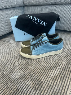 Lanvin blå  - Snygga sneakers från Lanvin med blå mocka på sidorna och glansig olivgrön tå och häl. Skorna har svarta skosnören och vit sula. Perfekt för dig som gillar en unik och stilren look med premiumkänsla.