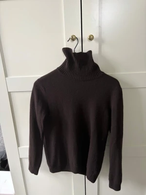 Mörkbrun polotröja Oscar Jacobson - Snygg mörkbrun stickad polotröja från Oscar Jacobson. Tröjan har hög ribbad krage och långa ärmar, perfekt för kyliga dagar. Tillverkad i mjukt material som känns skönt mot huden. Enkel och stilren design som passar till mycket.