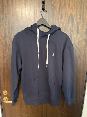 Mörkblå hoodie från Polo Ralph Lauren - Mörkblå hoodie från Polo Ralph Lauren med vit broderad logga på bröstet och vita dragsnören i huvan. Klassisk känguruficka framtill och ribbade muddar vid ärmslut och nederkant. Tillverkad i mjuk bomullsblandning för extra komfort.