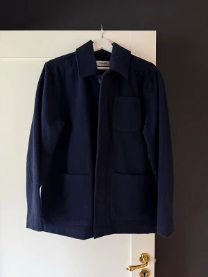 Ulljacka - Stilren mörkblå ulljacka. Rak passform med klassisk krage, tre stora utanpåfickor och dold knappslå framtill. Perfekt lager-på-lager-plagg för en avslappnad men snygg look.