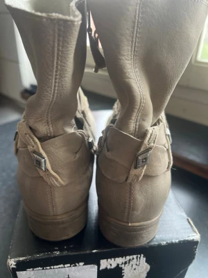 Beige stövletter med dekorativt spänne - Snygga beige stövletter i mjukt skinn med dekorativt band och metallspänne runt ankeln. Låg klack och rundad tå gör dem både stilrena och bekväma. Perfekta för dig som vill ha en enkel men trendig look.