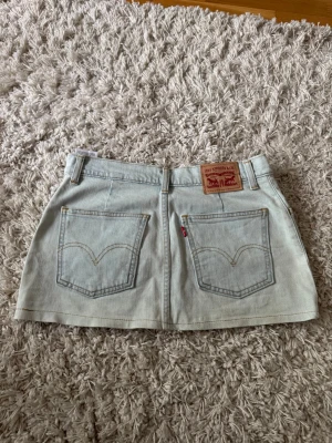 Ljusblå jeanskjol från Levi's - Snygg ljusblå jeanskjol från Levi's med klassiska bakfickor och knappstängning framtill. Kjolen har bälteshällor och en rak, kort passform. Perfekt för dig som gillar en avslappnad och trendig stil.