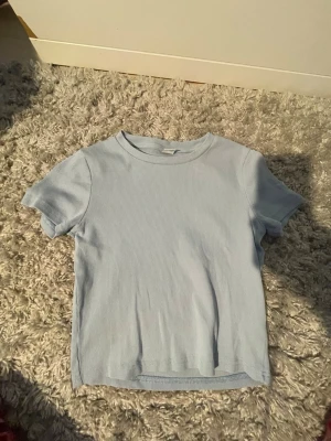 Ljusblå ribbad t-shirt - En ljusblå ribbad t-shirt med korta ärmar och rund halsringning. Toppen har en enkel och fin design som är lätt att matcha med olika outfits. 