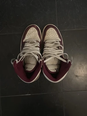 Nike Air Jordan 1 Bordeaux Highs - Snygga Nike Air Jordan 1 Highs i färgen Bordeaux och vitt. Skorna har klassisk siluett med högt skaft, vita snören och silverfärgad swoosh. Ovandelen är i läder med perforerad tåbox och detaljer i vinrött. Perfekta för dig som gillar streetwear och sneakerskultur.