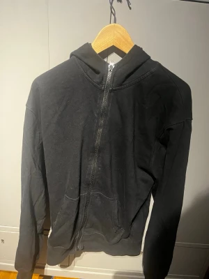 Svart Gasp zip med huva - Svart zip från Gasp med huva. Tröjan har tryck på ena ärmen och är perfekt för den som gymmar. Materialet känns mjukt och bekvämt. Storlek S men är oversized runt överkroppen.