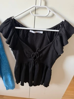 Svart blus med volang och knyt - Svart blus med volangdetaljer och knyt framtill. Toppen har en v-ringad urringning och är tillverkad i ett lätt, strukturerat tyg. Modellen är axelbandslös och har en luftig passform med rynkade detaljer nedtill.
