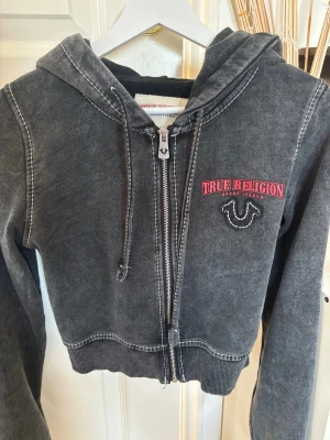 Svart hoodie från True Religion - Snygg svart zip-hoodie från True Religion med stor logga i rött och vitt både fram och bak. Hoodien har huva med dragsko, ribbade muddar och kontrastsömmar. Perfekt för en avslappnad streetstyle-look.