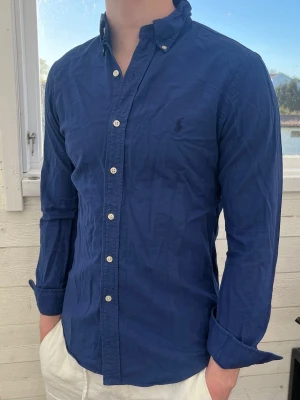 Marinblå skjorta från Ralph Lauren - Klassisk marinblå skjorta från Ralph Lauren med button-down krage och broderad logga på bröstet. Skjortan har långa ärmar, vita knappar och är tillverkad i mjuk bomull. Perfekt till jeans eller chinos för en clean och stilren look.