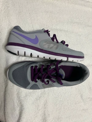 Grå och lila Nike sneakers - Snygga sneakers från Nike i grått med lila detaljer och lila snörning. Skorna har meshpaneler för extra andningsförmåga och en vit sula med lila kant. Perfekta för dig som gillar sportig stil och vill ha bekväma skor till vardagen.