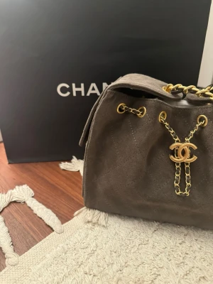 Chanel hobo 26  - Säljer en beige/grå  handväska från Chanel i mjuk mocka med klassiskt rutmönster. Väskan har guldfärgade kedjedetaljer och ikoniska CC-loggan framtill. Perfekt för dig som gillar lyxiga accessoarer med tidlös design. Sparsamt använd. Mycket bra skick. Kommer med låda, dustbag. 