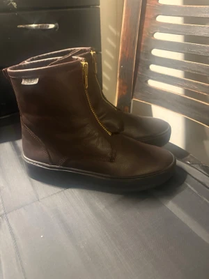 Bruna stövletter med gulddragkedja - Snygga bruna stövletter i läder med guldfärgad dragkedja framtill. Skorna har en stilren och enkel design med rund tå och låg sula, perfekta för dig som gillar en clean look. Passar till många olika outfits och är lätta att ta på tack vare dragkedjan.