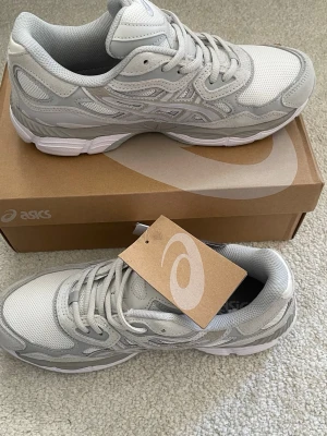 Gråa ASICS  - Snygga gråa ASICS sneakers med ovandel i mesh och detaljer i mocka och syntet. Skorna har klassisk ASICS-logga på sidan, vita sulor och grå snörning. Perfekta för dig som gillar stilrena och sportiga sneakers med modern vibe.