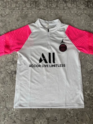 PSG x Jordan vit träningströja - Säljer en snygg vit träningströja från PSG x Jordan med svarta och rosa detaljer. Tröjan har halv dragkedja, rosa mönstrade ärmar och PSG-logga på bröstet. Materialet är lätt och andas, perfekt för träning eller fotboll.