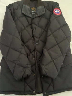 Svart dunjacka från Canada Goose - Säljer en svart dunjacka från Canada Goose med klassiskt rutmönster och hög krage med läderdetaljer. Jackan har tryckknappar framtill och den ikoniska loggan på ärmen. Perfekt för kalla vinterdagar.