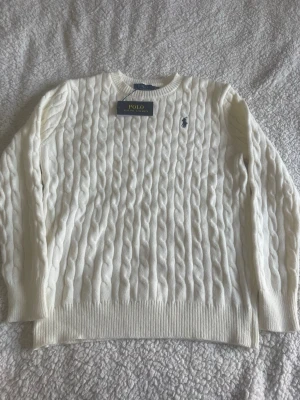 Vit kabelstickad tröja Polo Ralph Lauren - Kabelstickad vit tröja från Polo Ralph Lauren med klassisk rund halsringning och den ikoniska broderade loggan i marinblått på bröstet. Tröjan har ribbade muddar vid ärmslut och nederkant. Perfekt för en stilren och tidlös look. De står att tröjan är i storlek L men passar M