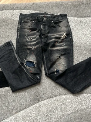 Svarta distressed jeans från Dsquared2 - Snygga svarta jeans från Dsquared2 med slitningar, hål och färgstänk för en edgy look. Jeansen har klassisk femficksmodell och patch med Dsquared2-logga bak. Tillverkade i denim med stretch för skön passform.