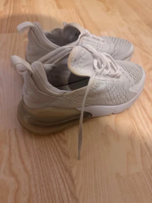 Nike Air Max 270 vita sneakers - Säljer ett par vita Nike Air Max 270 sneakers med stor synlig Air-bubbla i hälen och meshdetaljer på ovandelen. Skorna har snörning och en sportig, modern look med diskreta loggor på sidan och bak. Perfekta för dig som gillar streetwear och bekväma skor.