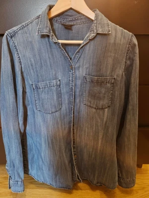 Blå jeansskjorta från Lindex - Snygg blå jeansskjorta från Lindex med två bröstfickor och klassisk krage. Skjortan har långa ärmar och knäppning framtill. Perfekt att styla lager på lager eller bära som den är.