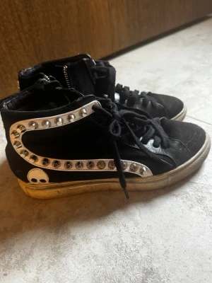 Svarta high top sneakers med döskallar - Unika svarta high top sneakers med vita detaljer, nitar och döskallemotiv på sidan och hälen. Skorna har snörning framtill och dragkedja på sidan. Materialmix av mocka och läderimitation. Perfekta för dig som gillar en edgy stil.