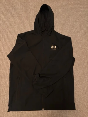 Svart vindjacka från Under Armour - Helt ny nästan oanvänd  svart vindjacka från Under Armour med dragkedja framtill och huva. Jackan har en minimalistisk design med logga på bröstet och två sidofickor med dragkedja. Tillverkad i lätt och slitstarkt material som passar perfekt för blåsiga dagar. 