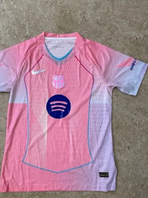 Barça x Nike rosa matchtröja - Säljer en rosa FC Barcelona matchtröja från Nike med klubbmärke, Nike-logga och Spotify-logga på bröstet. Tröjan har korta ärmar, blå och röda detaljer samt UNHCR-tryck på ryggen. Tillverkad i lätt och ventilerande funktionsmaterial.