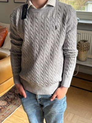 Grå kabelstickad tröja Polo Ralph Lauren - Snygg grå kabelstickad tröja från Polo Ralph Lauren med rund halsringning och broderad logga på bröstet. Tröjan har ribbade muddar och är perfekt att bära över en skjorta för en klassisk look.