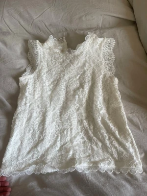 Vit spetsblus med volangdetaljer - Supersöt vit blus i spets med fina volangdetaljer vid ärmslut och hals. Ärmlös modell med vacker broderad spets och lätt v-ringad halsringning. Perfekt för dig som gillar romantisk och feminin stil.