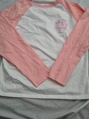 Chrome Hearts långärmad topp rosa/vit - Långärmad topp från Chrome Hearts med vita paneler och ljusrosa ärmar. Stort rosa tryck med logga och kors på ryggen samt mindre logga framtill. Skön bomullskvalitet och avslappnad passform, perfekt för en cool och avslappnad stil.  Nypris är 20 000.