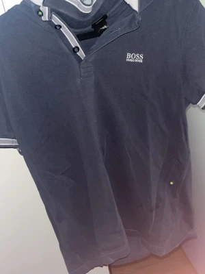 Mörkblå pikétröja från Hugo Boss - Snygg mörkblå pikétröja från Hugo Boss med vit logga på bröstet. Tröjan har krage med knappar och vita detaljer på krage och ärmslut. Regular fit och kortärmad, perfekt för en clean och stilren look.