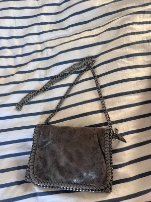 Grå axelväska med kedjedetaljer - Snygg grå axelväska från Stella McCartney med kedjerem och dekorativa kedjor runt kanterna. Väskan har en magnetisk knappstängning och innerfack med dragkedja. 