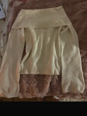 Off shoulder stickad tröja cremevit - Trendig off shoulder stickad tröja i cremevitt med bred ribbad kant upptill och vid ärmslut. Mjuk och skön kvalitet, perfekt för lager på lager. Långa ärmar och avslappnad passform ger en snygg siluett.