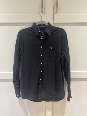 Svart skjorta från GANT, regular fit - Klassisk svart skjorta från GANT i regular fit med vita knappar och bröstficka med broderad logga. Skjortan är tillverkad i bomull och har button down-krage. Perfekt för en stilren och enkel look.