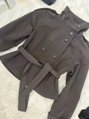 Kort brun trenchcoat med bälte - Snygg kort trenchcoat i mörkbrunt med dubbelknäppning och höga axlar. Jackan har ett matchande knytbälte i midjan och dekorativa axelklaffar. Tillverkad i ett slätt tyg, perfekt för lager-på-lager-stil. Finns liten missfärgning syns på bilder