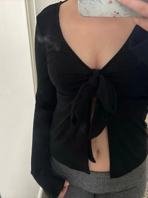 Svart långärmad topp med knyt - Trendig svart långärmad topp med djup v-ringning och knyt framtill. Perfekt att styla med höga jeans eller kjol. Mjuk och stretchig känsla. Säljer då den inte är min stil