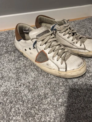 Philippe Model vita sneakers med bruna detaljer - Snygga vita sneakers från Philippe Model med bruna detaljer vid hälen och grå snörning. Skorna har en ikonisk sköldlogga på sidan och är tillverkade i läder med textil vid hälen. Perfekta för en trendig och avslappnad stil.