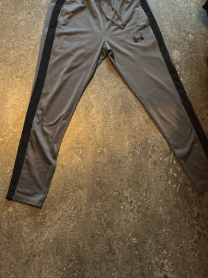 Grått träningsset från Under Armour - Säljer ett grått träningsset från Under Armour med svarta detaljer längs sidorna. Setet består av en zip-jacka med hög krage och matchande byxor med dragsko i midjan. Materialet är mjukt och stretchigt, perfekt för träning eller chill.