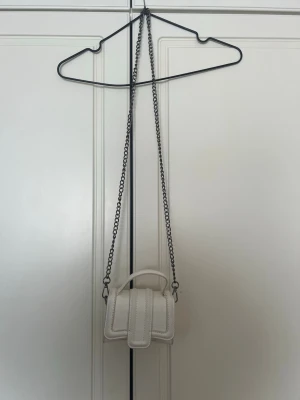 Vit väska med kjedja  - Superfin liten handväska i vitt med krokopräglat mönster och silverfärgad kedjerem. Väskan har ett stilrent handtag och en modern, boxig form. Perfekt accessoar för att lyfta din outfit.🤍